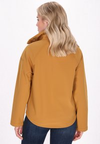 Žena s vlnit�ými blond vlasmi sa otáča, oblečená v horčicovo žltej bunde a tmavých džínsach, na svetlom pozadí.