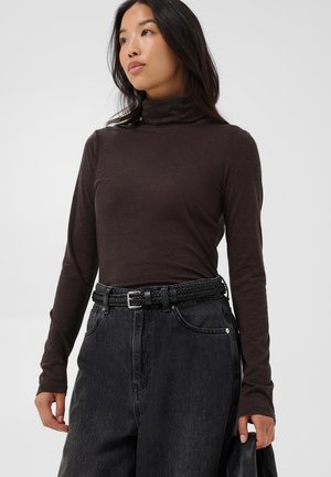 Jeune femme aux longs cheveux foncés porte un col roulé marron foncé et un jean noir taille haute avec une ceinture noire tissée, tenant un sac noir.
