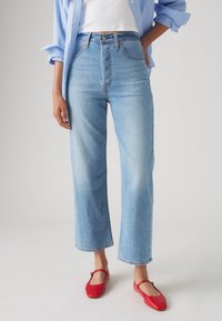 Femme portant un jean bleu clair droit, un haut blanc, une chemise bleue et des chaussures plates rouges Mary Jane, debout sur un fond uni.