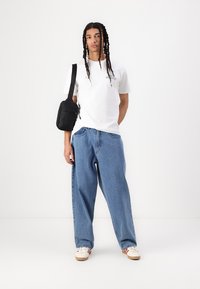 White cotton t-shirt with logo, baggy blue denim jeans, and white sneakers with red stripes. Black crossbody bag on left shoulder.