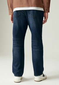 Vaqueros de mezclilla azul oscuro con un corte relajado, que cuentan con bolsillos traseros y detalles sutiles de desgastado en los muslos. Combinados con zapatillas blancas.