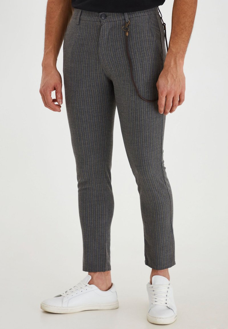 Grau gestreifte Hose mit schmalem Schnitt, mit Knopfverschluss und einer geschlungenen braunen Kette als Akzent. Getragen mit weißen Sneakers.