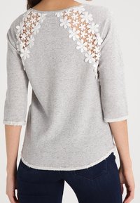 Top a rayas grises con mangas 3/4, que presenta detalles de encaje floral blanco en los hombros y un dobladillo deshilachado para un acabado texturizado.