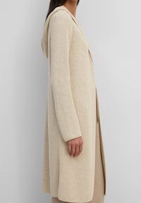 Cardigan beige lavorato a maglia con cappuccio, maniche lunghe e design aperto. Tessuto testurizzato con una vestibilità ampia e dettagli a coste sulle maniche.