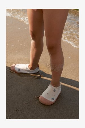 Hvite vannsko uten snøring med rosa såler, dekorert med små blomster. Skoene bæres på sandete føtter som delvis er våte fra stranden.