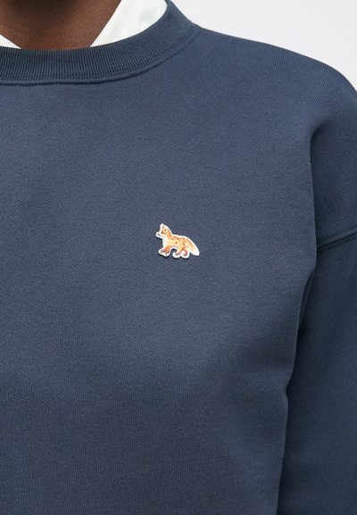 Nokrāsota jāntī zila kapučjakete ar elastīgu apkakli un mazu izšūtu oranžu lapsas emblemu uz krūtīm, izgatavota no teksturēta auduma.