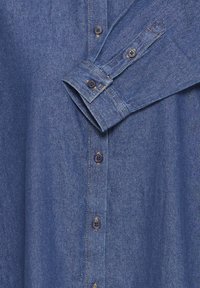 Chemise en denim avec un tissu bleu, dotée d'un devant à boutons, de manches longues avec poignets boutonnés et de boutons foncés. Coutures visibles le long des coutures.