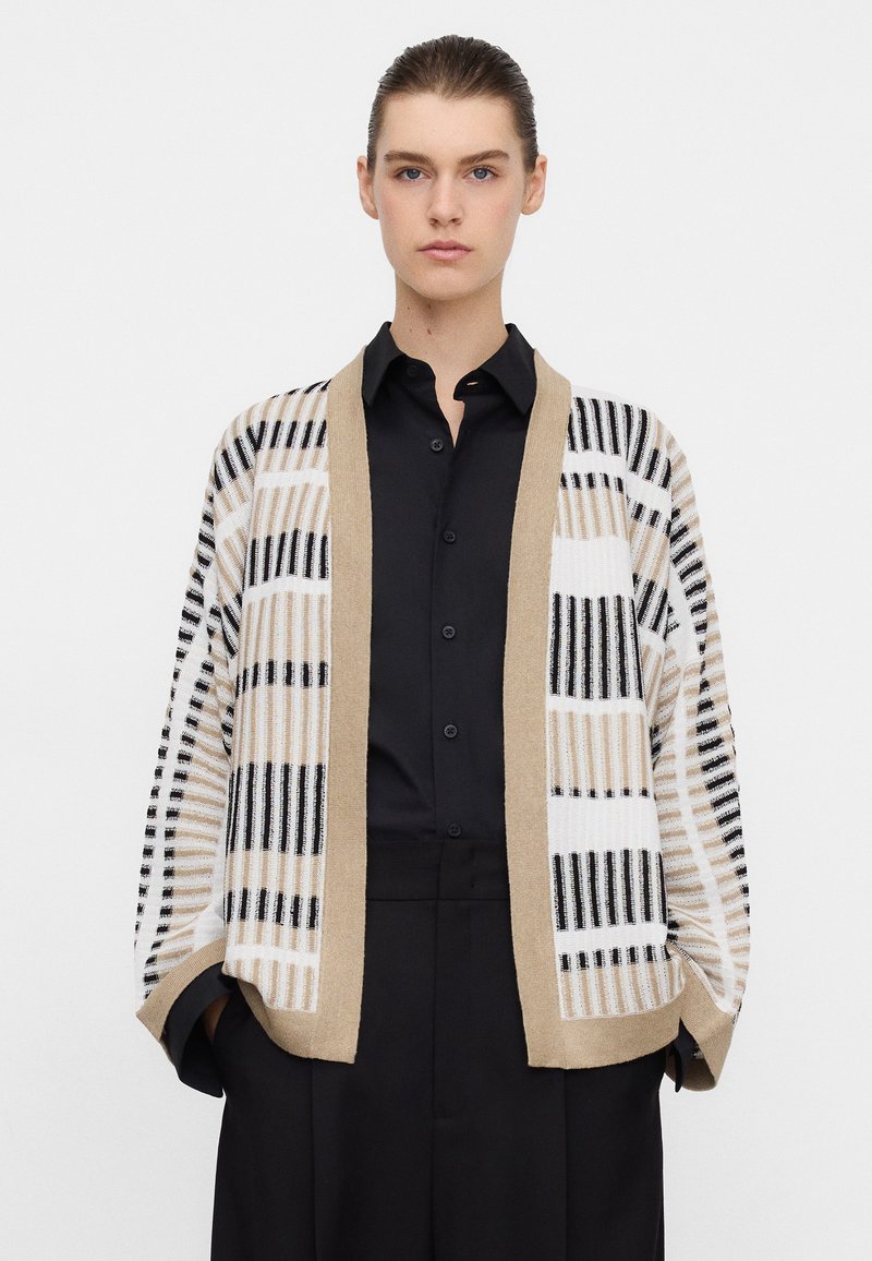 Young person with slicked-back hair wears a beige, white, and black striped open-front cardigan over a black button-up shirt and black pants.