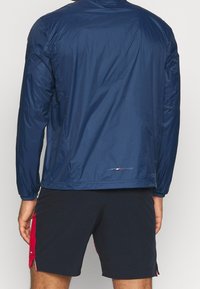 Chaqueta ligera de color azul marino con una textura suave, mangas largas y corte recto. Presenta un logo en el dobladillo inferior.