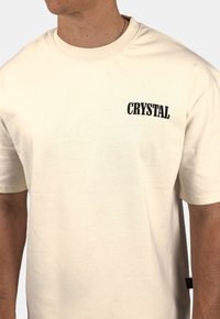 Krémové bavlněné tričko s krátkým rukávem a zaobleným výstřihem, na levé části hrudi s výrazným černým nápisem "CRYSTAL".
