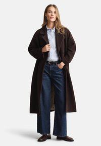 Dlouhý hnědý vlněný kabát se širokými klopami a zapínáním na knoflíky, přes světle modrou košili, doplněný širokými džíny a tmavě hnědými botami.