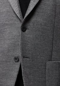 Blazer grigio a costine con rever a prefetto, due bottoni neri e una tasca sul lato destro, indossato sopra un indumento nero.