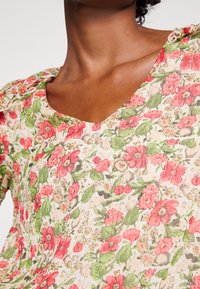 Gros plan d'une personne portant une robe à col en V à motif floral rouge, rose et vert sur un fond beige clair.