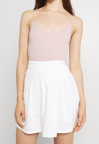 Světle růžový žebrovaný body s tenkými ramínky, doplněný bílou plisovanou sukní s přiléhavým pasem. Jednoduchý, čistý design.