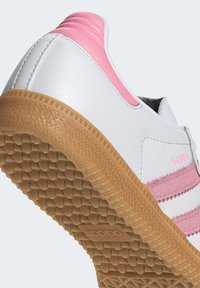 Biela kožená teniska s ružovými akcentmi a tromi pruhmi po boku, s textúrovanou podrážkou z gumy v prírodnej farbe a zaoblenou špičkou.