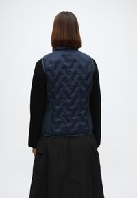 Ναυτικ�ό γυαλισμένο γιλέκο με ψηλό γιακά, διαθέτοντας υφή και πλαϊνές τσέπες, συνδυασμένο με ένα μαύρο ρούχο με μακριά μανίκια από κάτω.