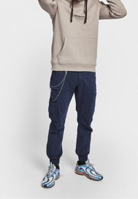 Beige hoodie with drawstrings and a front pocket, paired with navy cargo pants featuring side pockets and a chain accent, worn with multicolored sneakers.