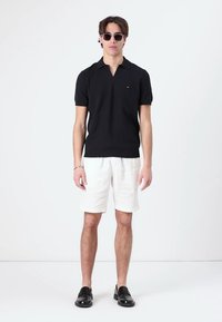 Schwarzes gestricktes Polohemd mit kurzen Ärmeln, weiße Leinen-Shorts, schwarze Schuhe und Sonnenbrille, vor einem schlichten weißen Hintergrund stehend.