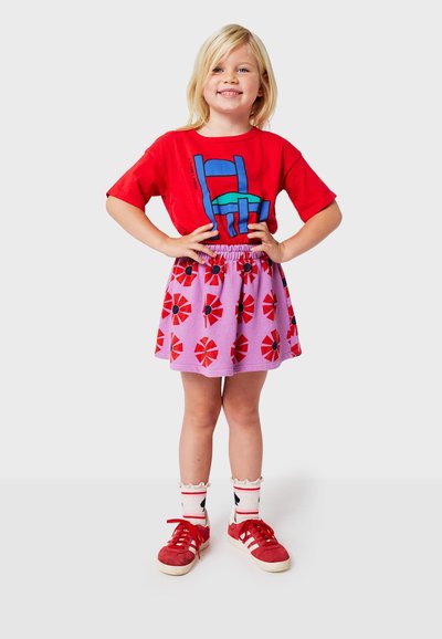 Rød grafik t-shirt med blå og grøn design, parret med en lilla nederdel med røde og sorte blomstermønstre. Røde sneakers og hvide sokker.