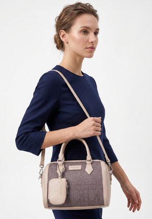 Frau in marineblauem Kleid hält eine beige und taupefarbene Handtasche mit abnehmbarem Täschchen vor einfachem hellem Hintergrund.