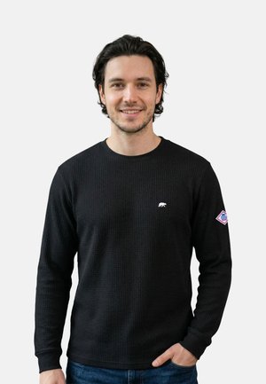 Lächelnder Mann mit dunklem Haar, der ein schwarzes, langärmeliges, strukturiertes Hemd mit kleinem weißem Logo auf der Brust und einem Aufnäher am Ärmel trägt.