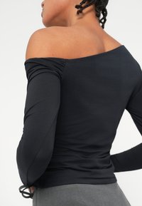 Crni top bez ramena s dugim rukavima, koji ima pripijen dizajn i skupljanje na ramenima, izrađen od glatkog, rastezljivog materijala.