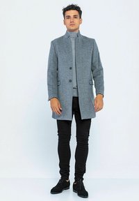 Grahamelierter Wollmischmantel mit hohem Kragen, Frontknöpfen und seitlichen Taschen, getragen über einem hellgrauen Rollkragenpullover und schwarzen Slim-Fit-Hosen.