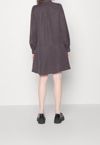 Vestito grigio con maniche lunghe e colletto sagomato, caratterizzato da un design fluido e dettagli di bottoni sui polsini, abbinato a scarpe nere.