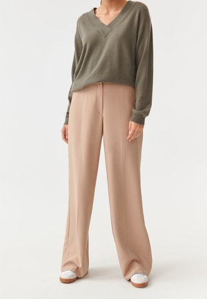 Pull tricot surdimensionné vert olive avec un col en V, associé à un pantalon beige à jambes larges. Chaussures blanches avec des accents marron. Textures lisses.