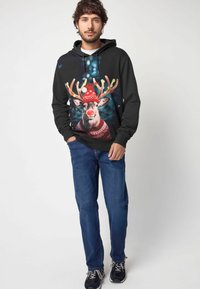 Sudadera negra con un gráfico de un reno con cuernos y un gorro festivo rojo. Combinada con vaqueros azules y zapatillas negras.
