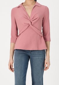 Rosa Langarmoberteil mit Kragen, das ein verdrehtes Frontdesign und einen ausgestellten Saum aufweist. Das Material fühlt sich weich an; mittlere Gewichtung und leicht strukturiert.