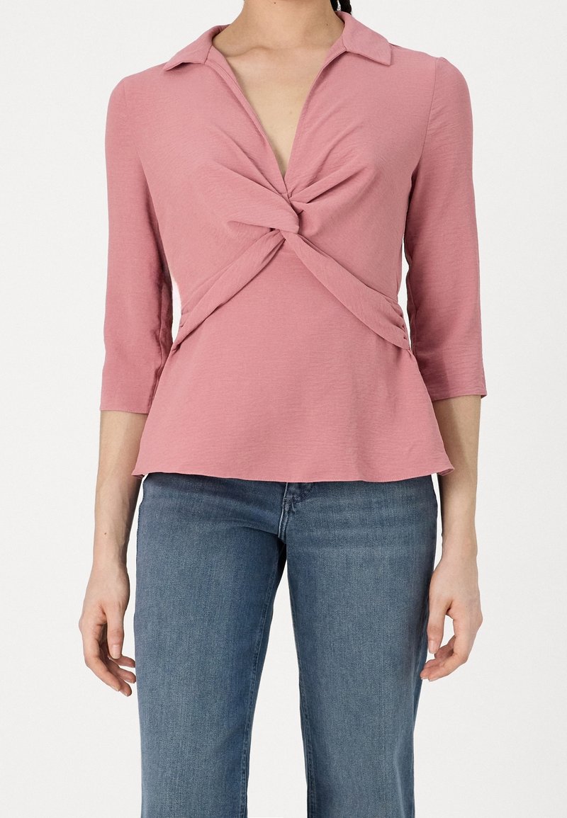 Rosa Langarmoberteil mit Kragen, das ein verdrehtes Frontdesign und einen ausgestellten Saum aufweist. Das Material fühlt sich weich an; mittlere Gewichtung und leicht strukturiert.