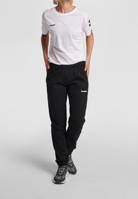 Weißes Baumwoll-T-Shirt mit schwarzem "hummel"-Logo; schwarze Jogginghose mit elastischem Bund und passendem Logo. Schwarze Sportschuhe mit grauen Akzenten.