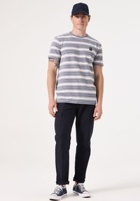 Camiseta de rayas de manga corta en azul marino y blanco, combinada con pantalones cargo en azul marino. Zapatillas blancas acentúan el conjunto. La gorra tiene un logo oscuro.