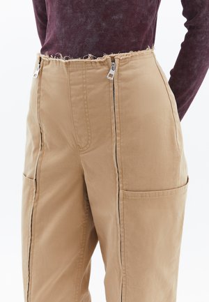 Pantalons cargo beiges avec de grandes poches latérales, des zips à l'avant et une taille effilochée ; fabriqués en tissu léger avec une texture lisse.
