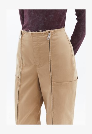 Pantalones de estilo cargo beige con grandes bolsillos laterales, cremalleras frontales y una cinturilla deshilachada; fabricados en un tejido ligero con una textura suave.