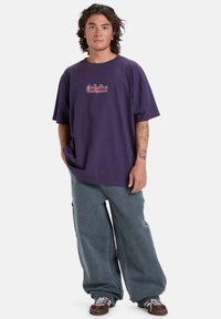 Lilla bomulds T-shirt med løs pasform, med grafisk broderi på brystet. Parret med lyseblå baggy jeans og mørke sko.