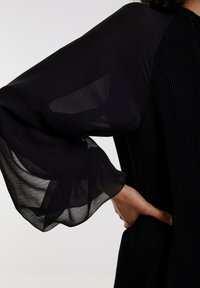 Zwarte jurk met doorschijnende, vloeiende mouwen en een gestructureerd, geplisseerd lijf. Het materiaal is lichtgewicht, wat beweging aan het ontwerp toevoegt.