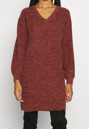 Person trägt ein langärmliges, rostrotes Strickkleid mit V-Ausschnitt und schwarze Hosen, steht vor einem schlichten hellen Hintergrund.