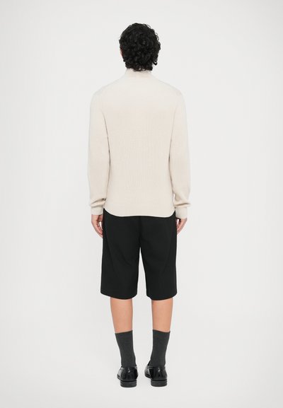 Μπεζ πλεκτό πουλόβερ με ριγέ ύφανση και μακριά μανίκια, σε συνδυασμό με μαύρο σορτς μέχρι το γόνατο. Φορεμένο με γκρι κάλτσες και μαύρα παπούτσια. Απλός σχεδιασμός, χωρίς σχέδια.