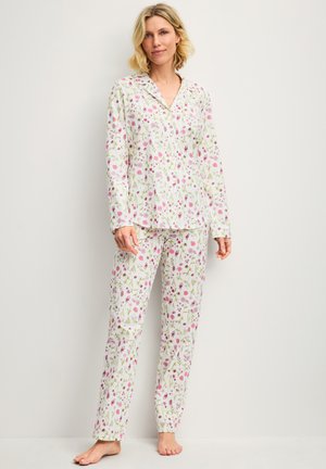 Femme debout pieds nus portant un ensemble de pyjama blanc à manches longues avec un motif floral rose et vert, contre un mur clair uni.