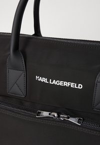Zwarte nylon draagtas met twee handvatten, een ritsluiting en witte logo-tekst. Bevat kenmerkende stiksels en een gladde textuur.