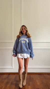 Maglione oversized azzurro chiaro con testo bianco "WRSTBVR 030", indossato sopra una gonna bianca a balze e stivali da cowboy beige, su un pavimento di legno.