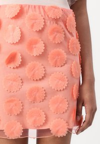 Gros plan sur une jupe rose corail avec des appliques florales superposées et un voile transparent, portée par une personne avec un bras et une main visibles.