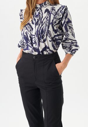 Mujer con blusa plisada de manga larga con patrón abstracto en azul marino y blanco, metida en pantalones negros de talle alto, con las manos en los bolsillos.