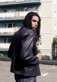 Black leather jacket with a smooth texture, featuring a classic design, long sleeves, and minimalistic detailing. Model stands outdoors.