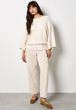 Beige oversized Pullover aus Wollmischung mit gerippten Bündchen, über einem hellbeigen, figurbetonten Oberteil getragen. Kombiniert mit cremefarbenen Hosen und braunen Ballerinas.