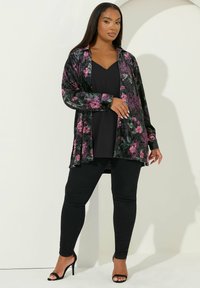 Cardigan in velluto nero con motivi floreali rosa e verdi, indossato sopra una blusa nera e abbinato a jeans skinny neri e tacchi.