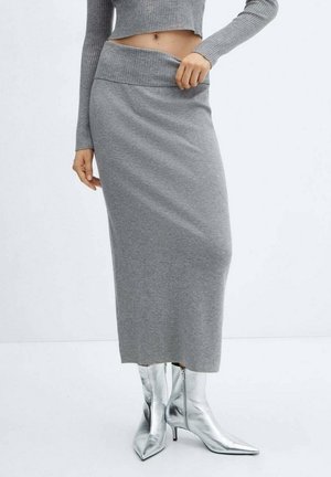 Kvinde iført en grå strikket midinederdel med høj talje, matchende grå kort sweater og sølvfarvede spidse hælede støvler står mod en hvid baggrund.