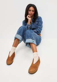 Bruine suède instappers met een V-vormige uitsnijding, gedragen met crèmekleurige geribde sokken en opgerolde blauwe denim.